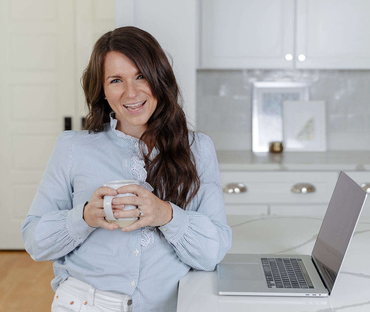 Personal brand photo used in a service page design showing a female entrepreneur standing in a bright kitchen workspace holding a coffee mug next to a laptop on a marble countertop.